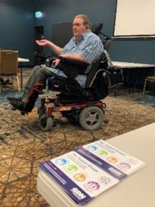 A man in a wheel chair at the front of a room talking.