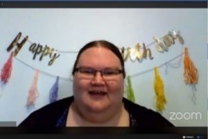 A screen shot of a Zoom call screen with a woman with a dark brown pulled back hair, wearing glasses talking.
