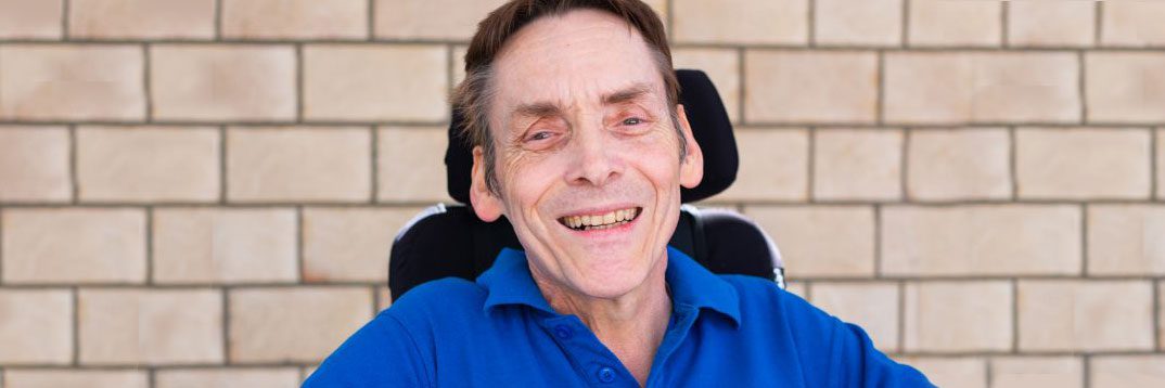 QDN board member Peter Tully is seated in a wheelchair wearing a blue polo and smiling at the camera.