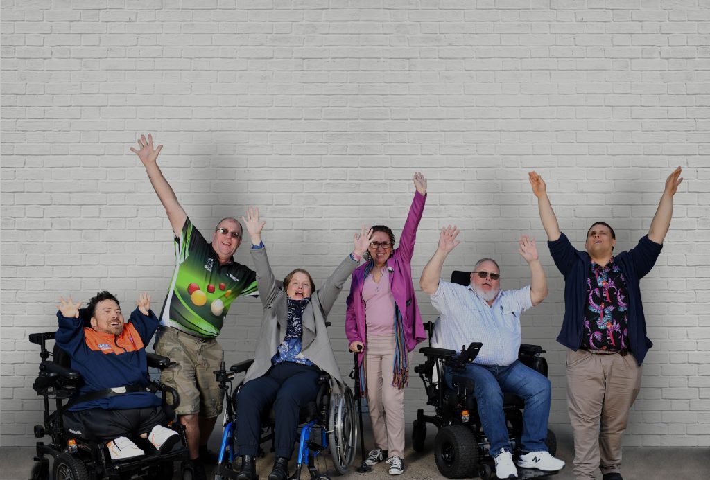3 people are standing, 2 men and 1 woman, and 3 people in wheelchairs, 2 men and 1 woman, are all raising their arms in the air. They are standing in front of a white bricked wall.