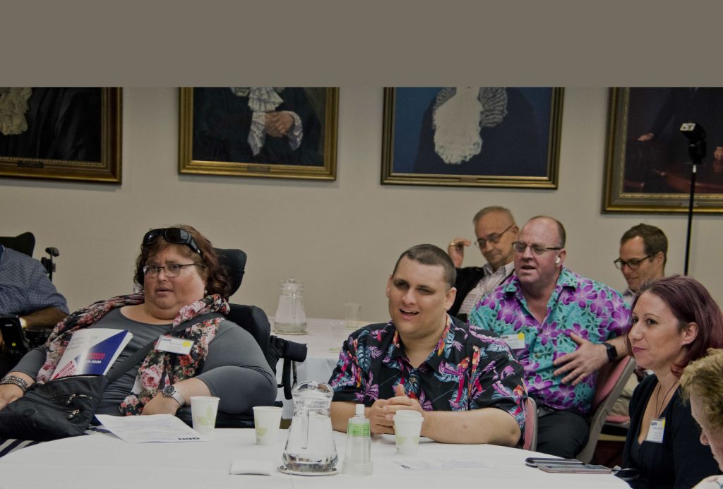 A lady in a wheelchair sitting at a table and a man in a colourful shirt is sitting next to her, there are other men and women sitting around them. There are glasses of water in front of them and papers on the table.