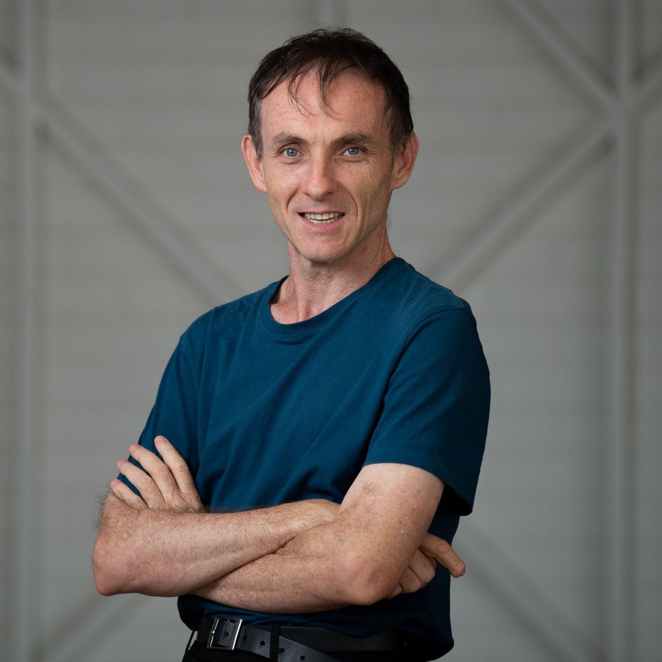 A man with his arms crossed wearing a teal tshirt and looking at the camera smiling.