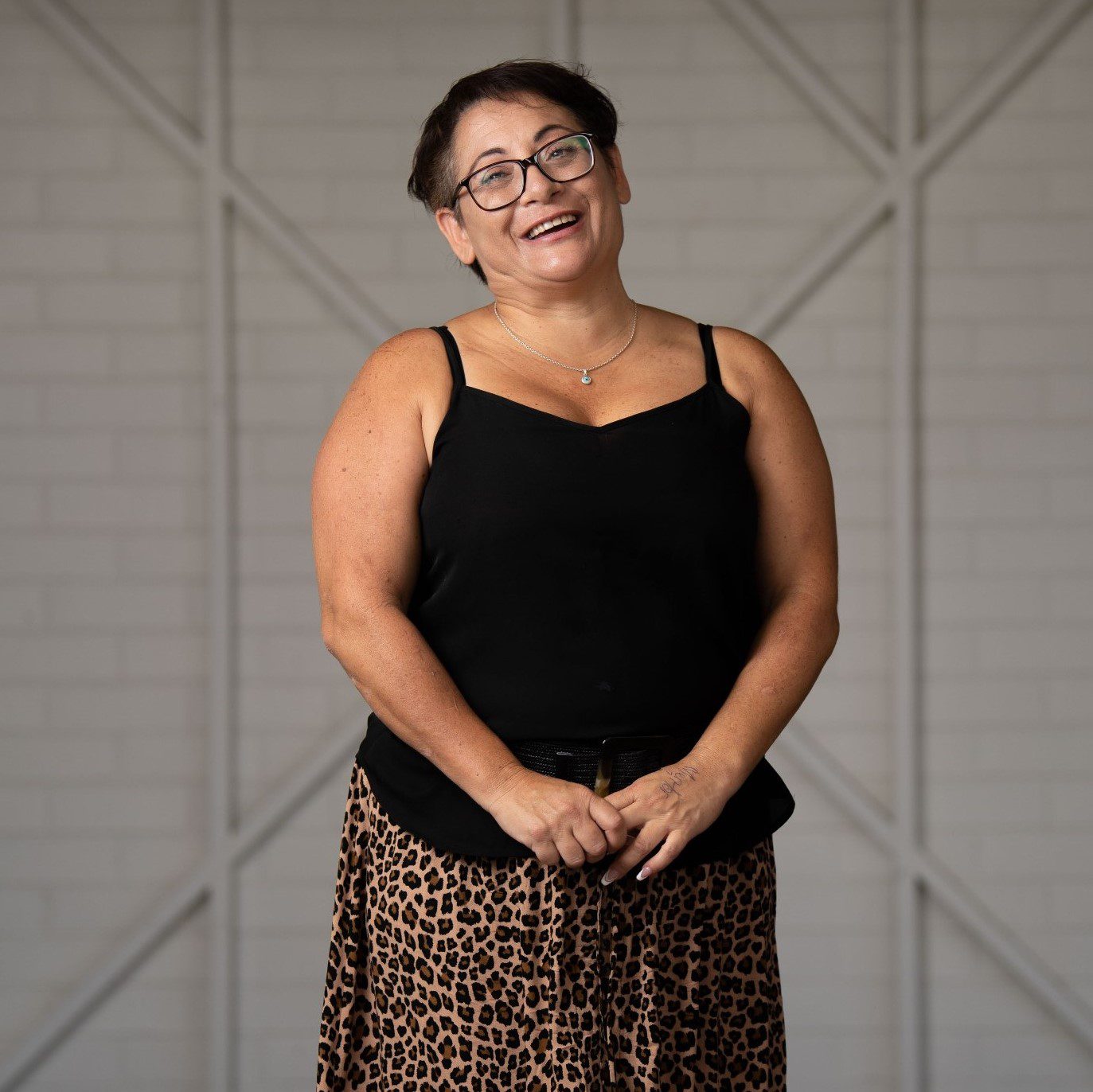 A lady wearing a black singlet and patterened skirt, she has short dark brown hair and is wearing glasses and smiling at the camera.