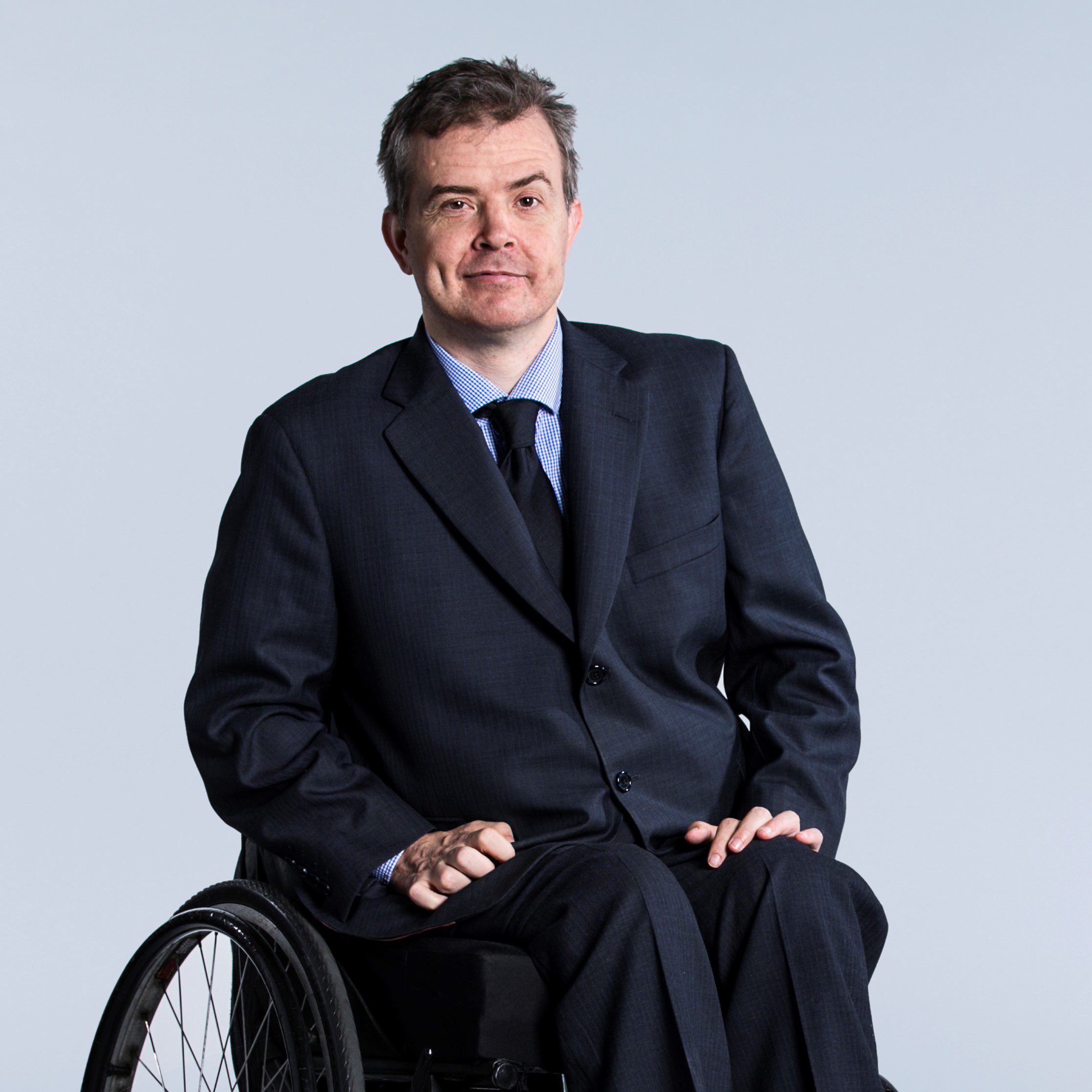 A man in a dark suit with a light blue business shirt and dark tie looking at the camera with his hands in his lap.
