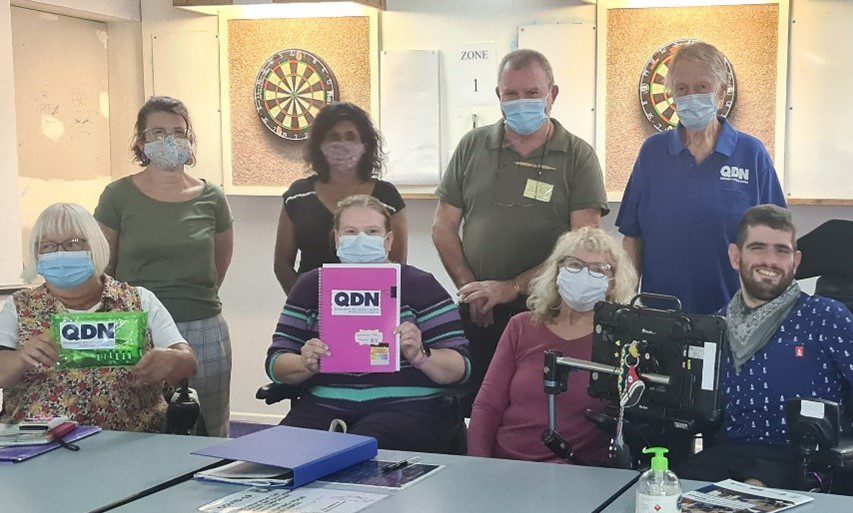 Four people standing behind another four people sitting at a table holding QDN resources. All wearing masks.