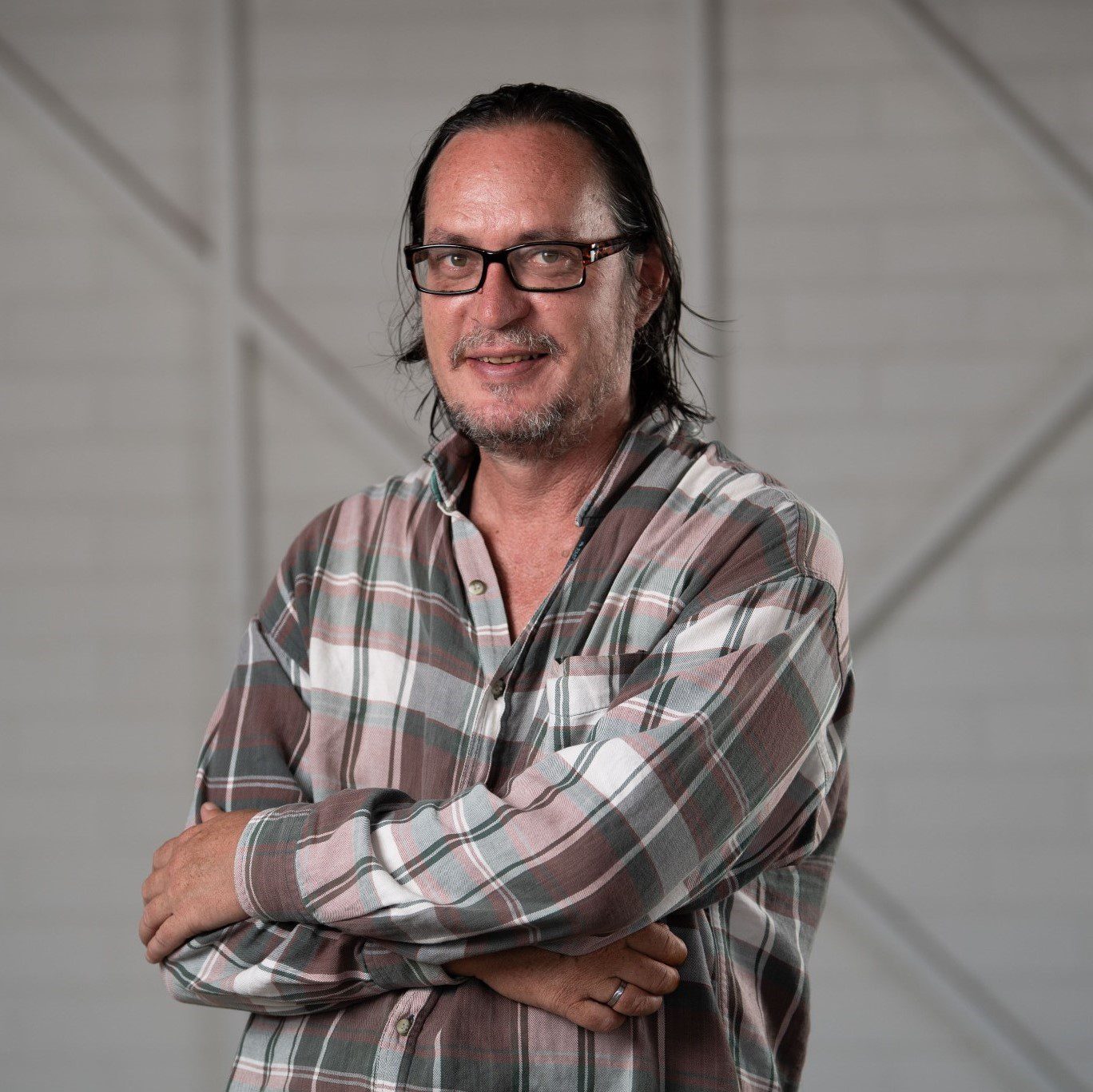 A man with shoulder length brown her and wearing glasses and a long sleeved checked shirt, he stands with his arms crossed smiling at the camera.