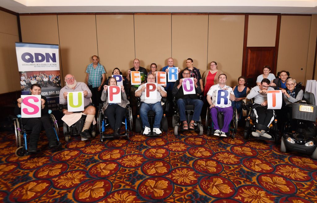 A group of people in wheelchairs and a group of people standing behind them are holding up colourful letters that spell Peer Support.