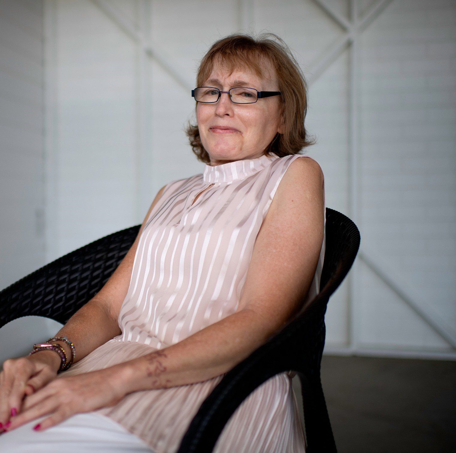 A lady sitting looking at the camera and smiling, she is wearing a light pink dressy shirt and white pants.
