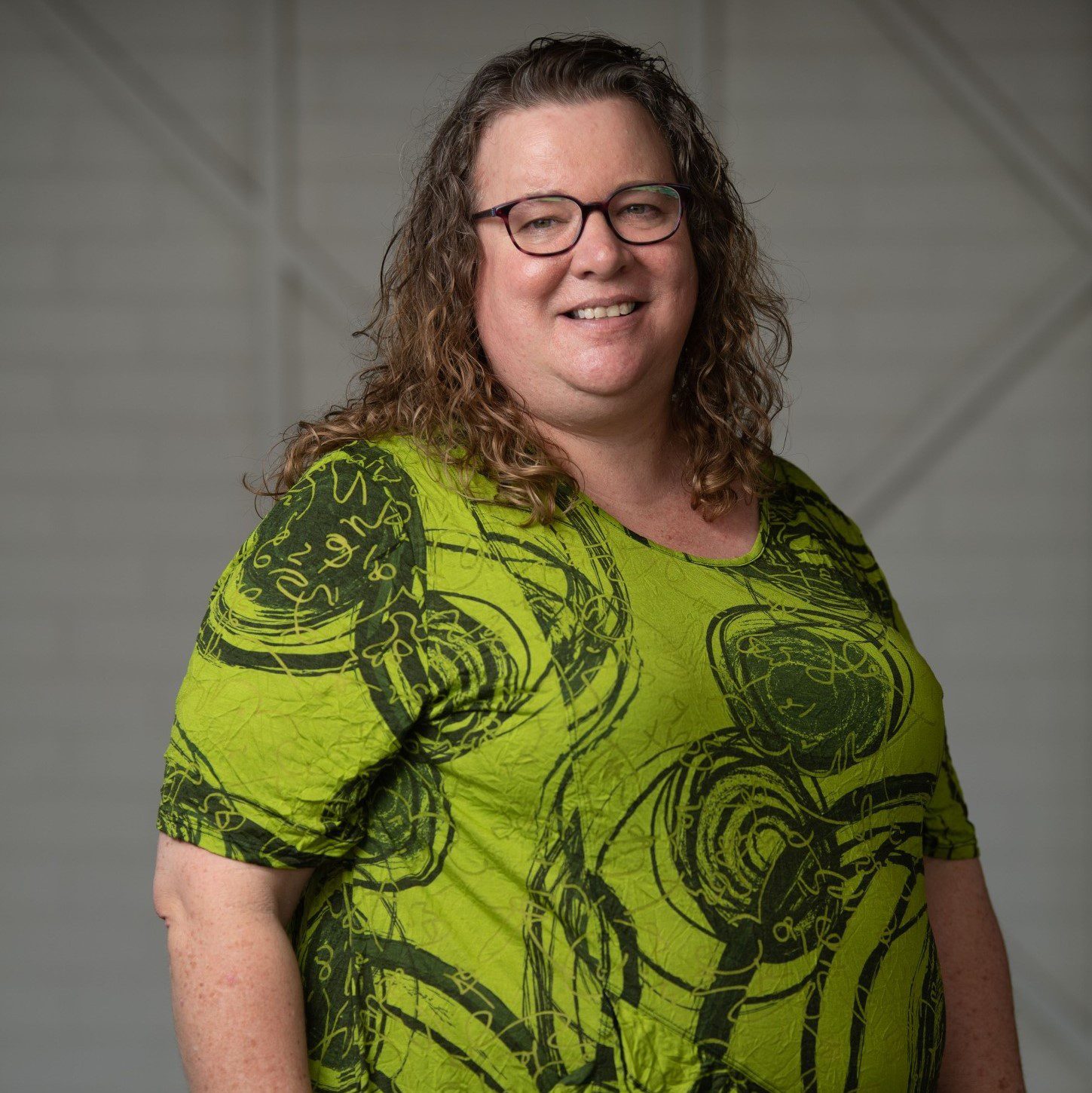 A lady with long light brown curly hair, wearing a green patterned shirt and glasses, looking at the camera smiling.