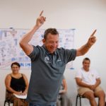 A man with his arms raised and pointing fingers upwards, standing in a room where people are sitting.