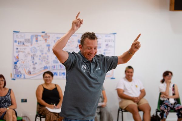 A man with his arms raised and pointing fingers upwards, standing in a room where people are sitting.