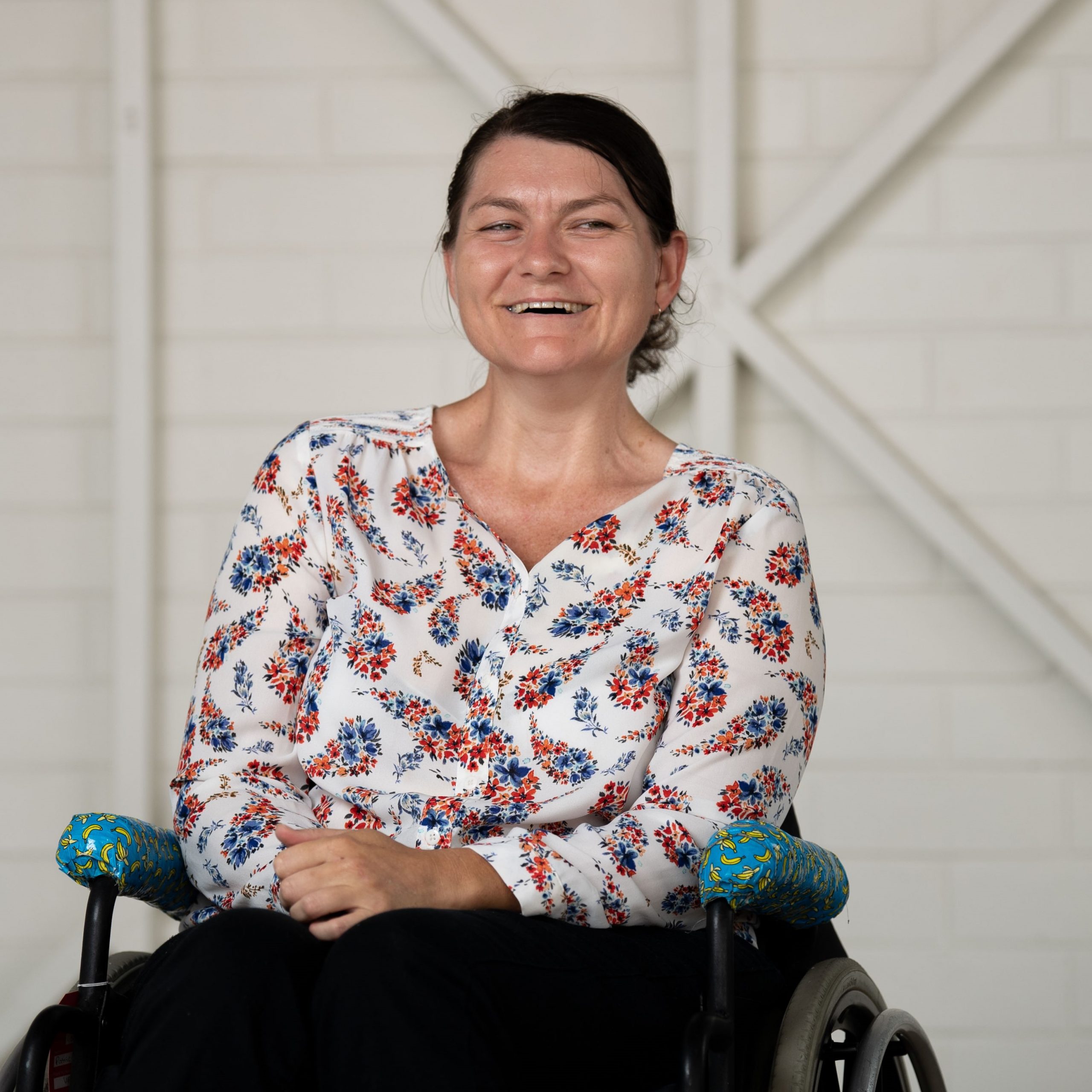 A lady with dark brown hair pulled back, wearing a white long sleeved shirt with colourful floral patterns and black pants. She is sitting with her hands in her lamp and smiling at the camera.