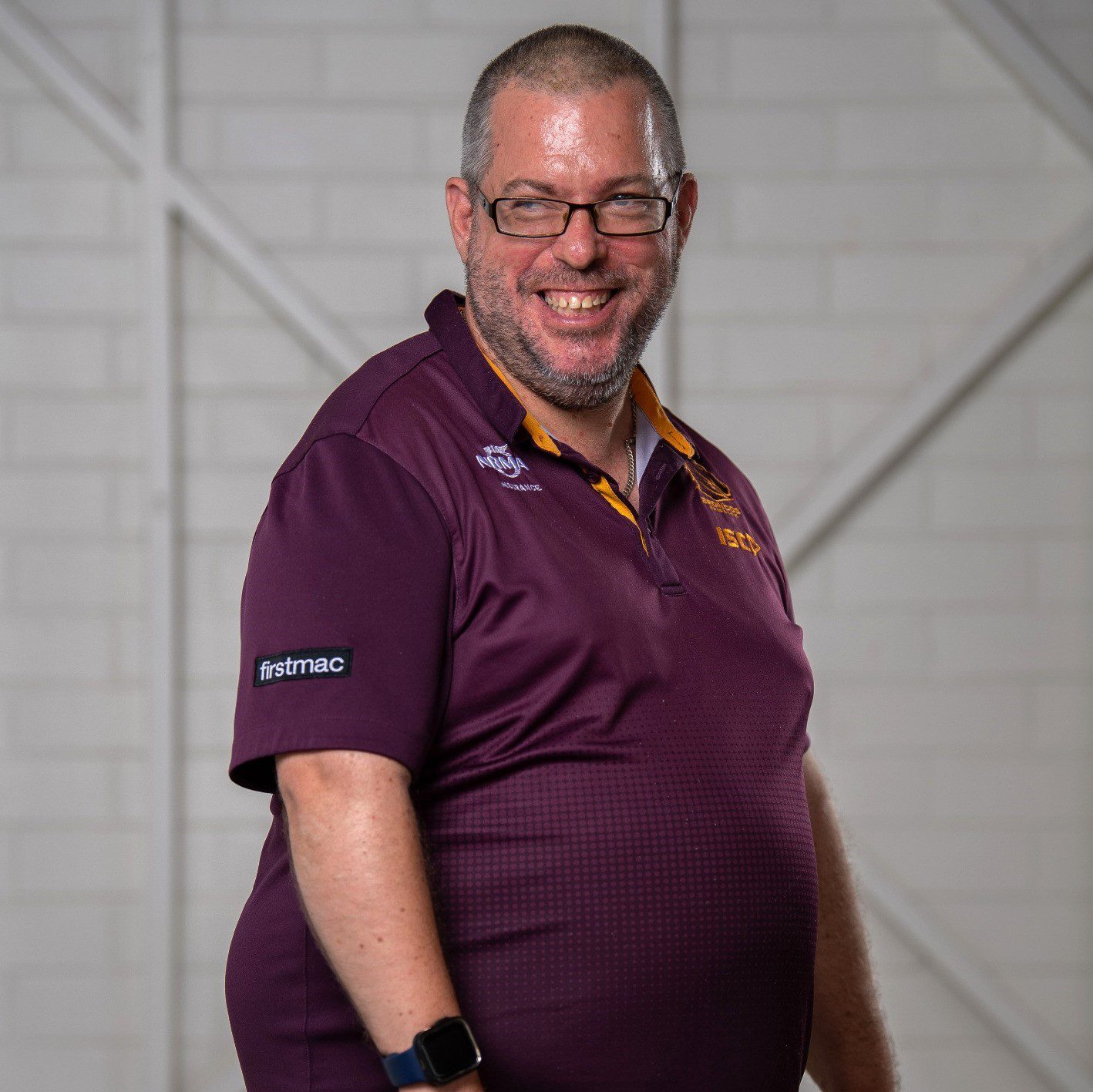 A man wearing a marron polo and glasses standing slightly to the side and smiling into the camera.