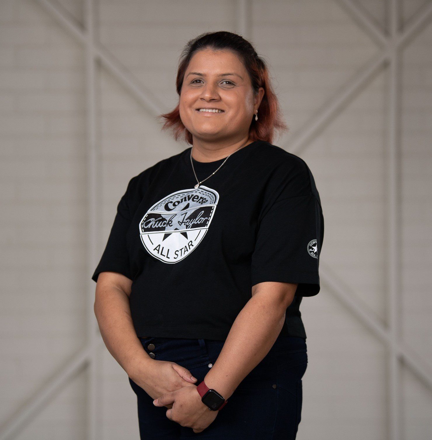 A woman in a black shirt and black pants, stands on her own in front of a white wooden wall. She is smiling and looking at the camera.