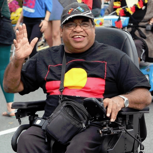 A photo of Uncle Willie Prince who is smiling and waving at the camera. He appears to be at an event, there are many people in the background behind him. He is sitting in a wheelchair, wearing a black cap and glasses. He is wearing a black t shirt with the Aboriginal flag, his hat has the Aboriginal flag and the Torres Strait Islands flag.