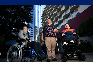A lady in a wheel chair next to a man holding a white cane and another man in a wheel chair next to him.