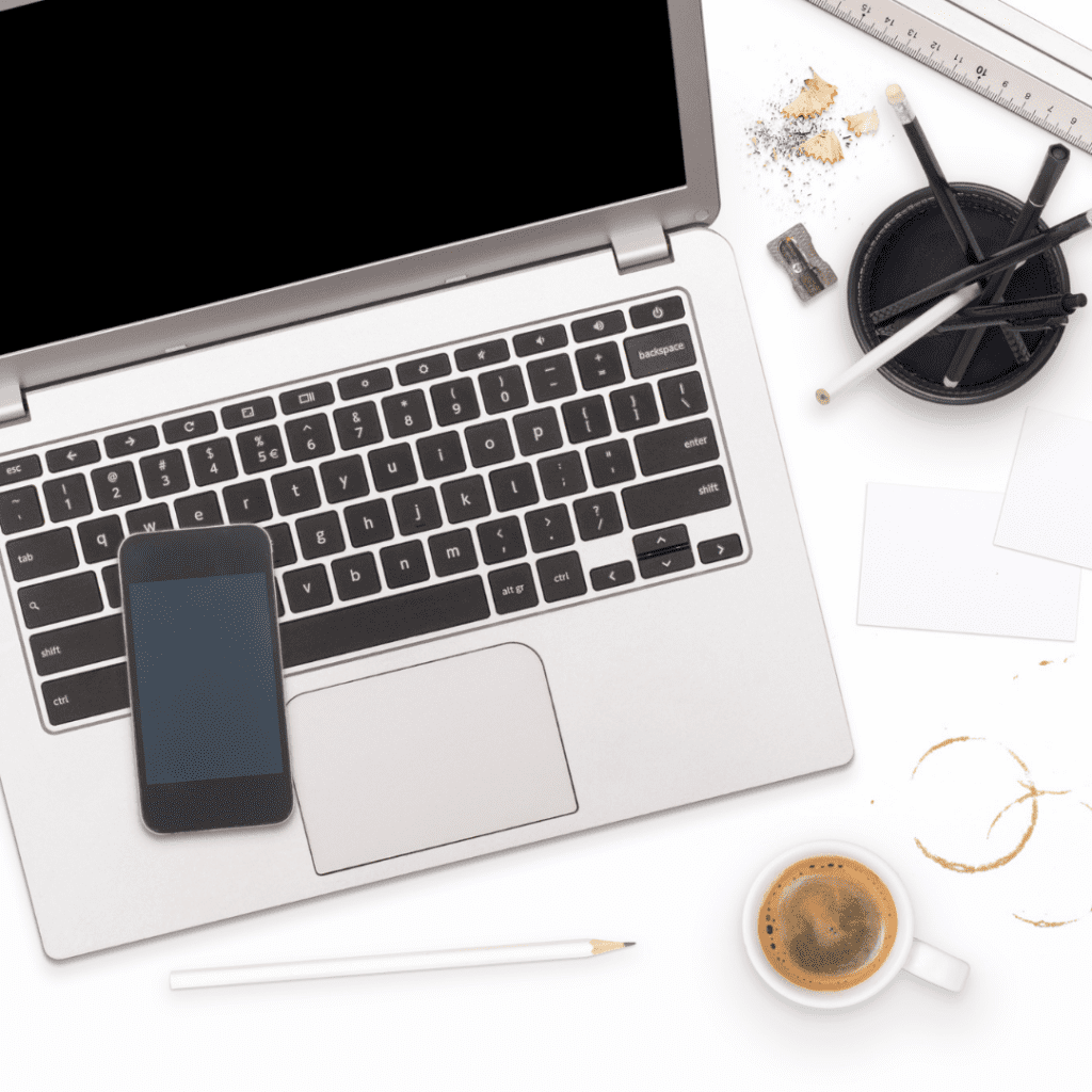 A photo of a laptop with a phone sitting on the keyboard, there are pencils in a holder and a coffee mug with coffee in it beside the laptop.