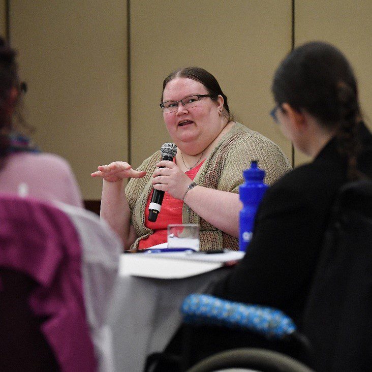 Foreground is out of focus and looking throw people seated and a woman holding a microphone is in focus. She is wearing a bright red shirt and light brown cardigan and wearing glasses, talking to the group.