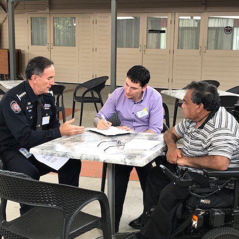 Three men sitting around a table, one in a emergency services uniform. One man taking notes and the man in the uniform is explaining something.