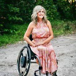 A woman with long blonde hair, sitting in a wheelchair wearing a pink long dress.
