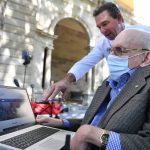 Two men outside. There is a man sitting in a wheelchair. He has a laptop on a table in front of him. Another man is standing and looking over his shoulder, pointing to the laptop screen.