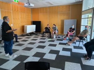 6 people in a room in chairs. They are all looking at a video screen.
