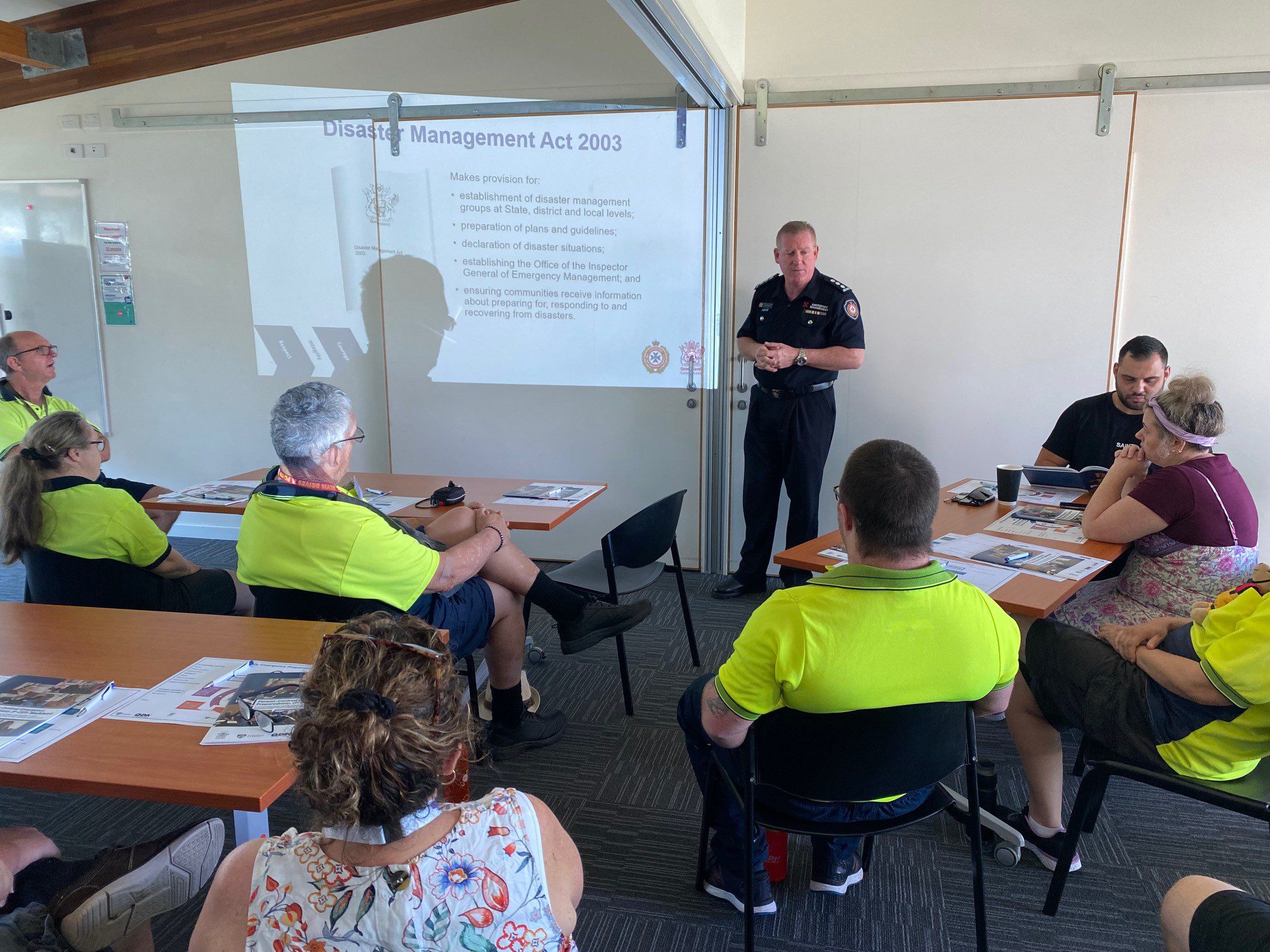 A group of people sitting behind tables at a workshop watching a man at the front of the room present. The man is wearing an emergency services uniform. 