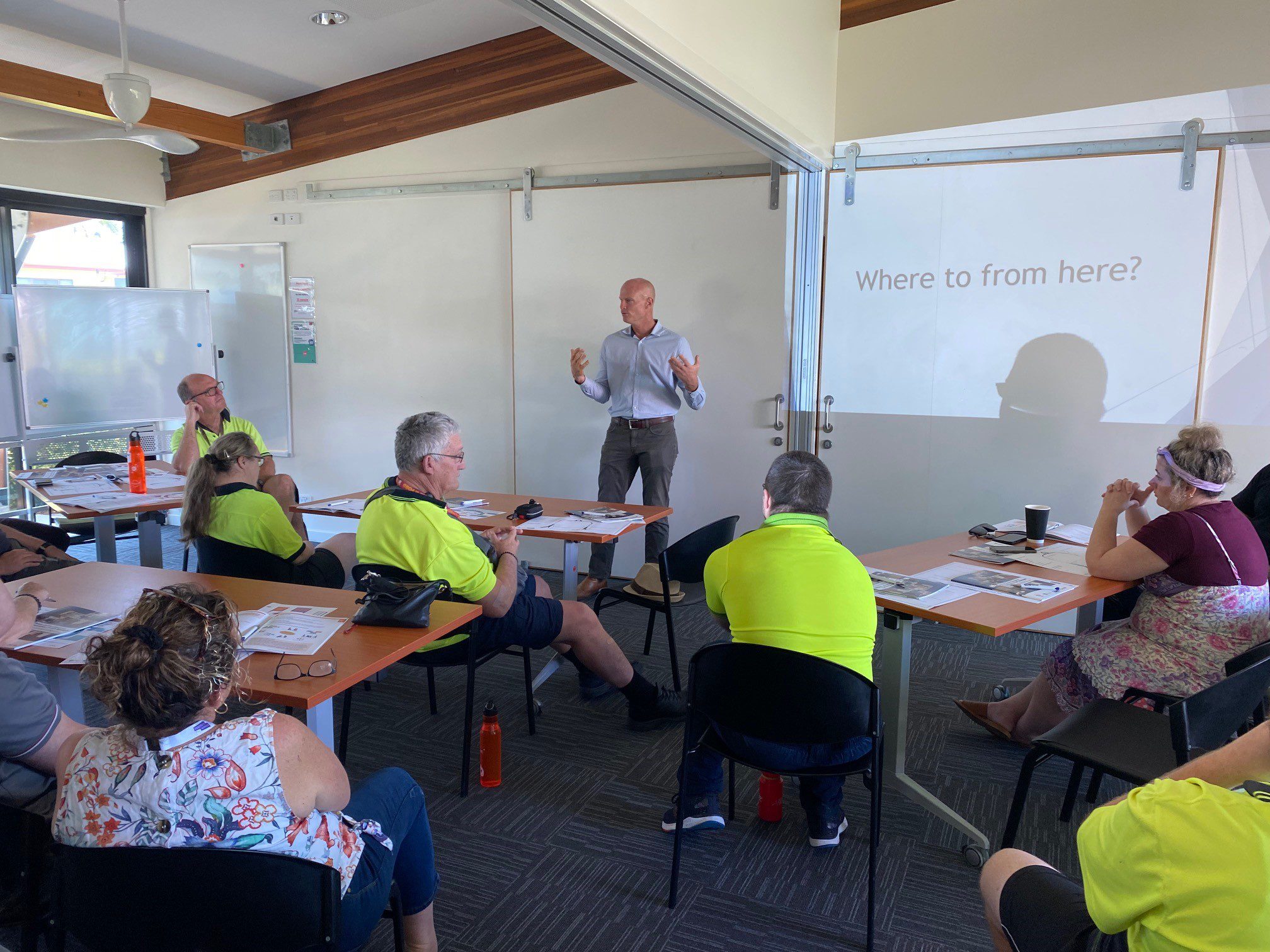 A group of people sitting behind desks facing a man speaking at the front of the room. The speaker is a tall man wearing a long sleeved buttoned up business shirt and slacks. A lot of the workshop participants are wearing high vis shirts. 