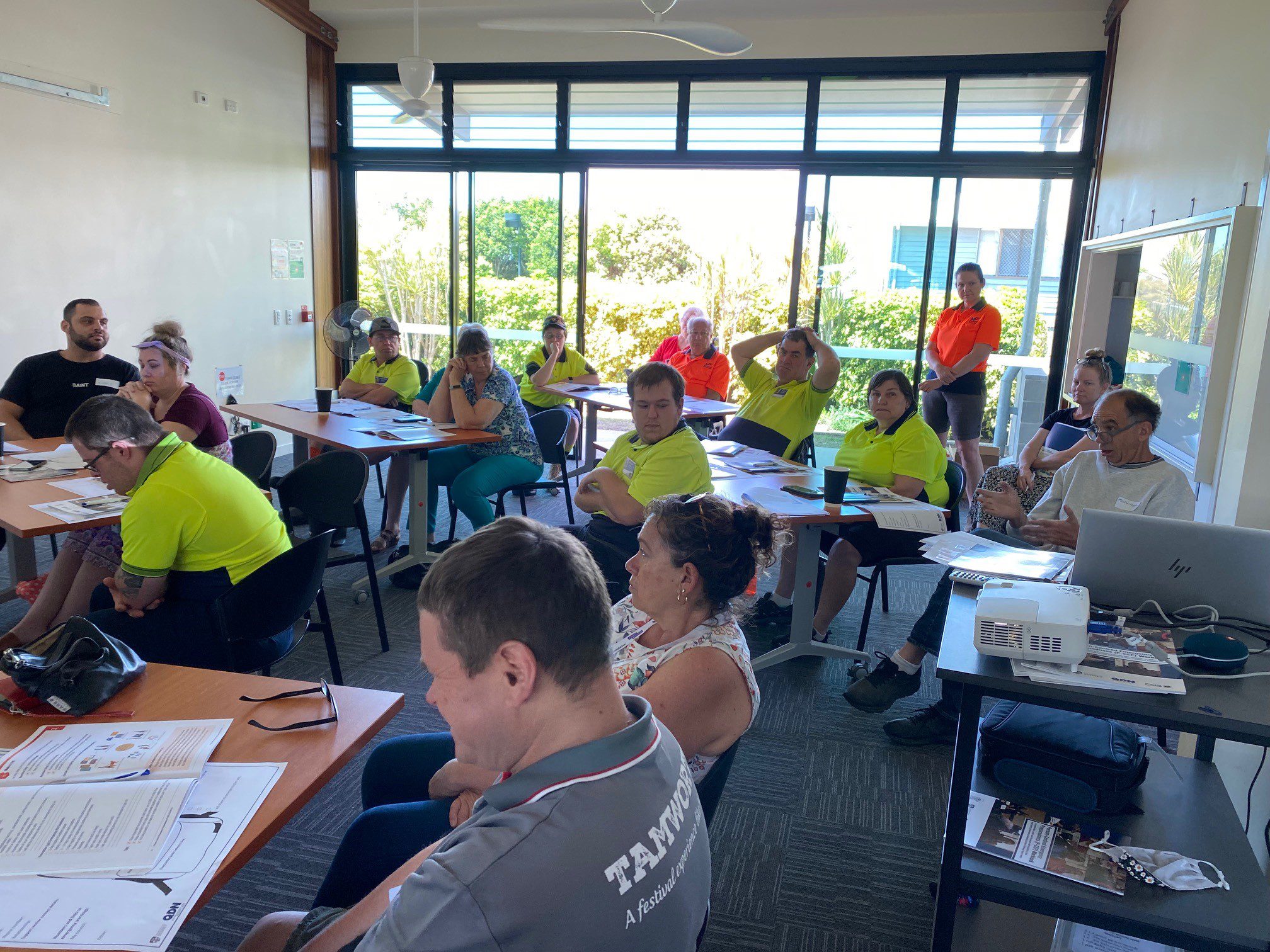 A group of people sitting behind desks at a workshop. Most of them are wear high vis fluro yellow polos. 
