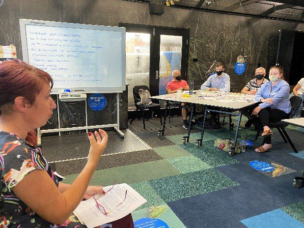 A lady with red hair is presenting to a group of people sitting at tables and they are all wearing masks. 