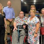 A group of 5 people standing and 1 sitting in a wheelchair. They are all looking at the camera smiling.