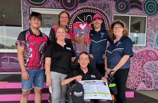 A group of people standing and one man in a wheelchair. They are in front of a wall with indigenous art on it and two of them are holding up a jersey with an indigenous design. 
