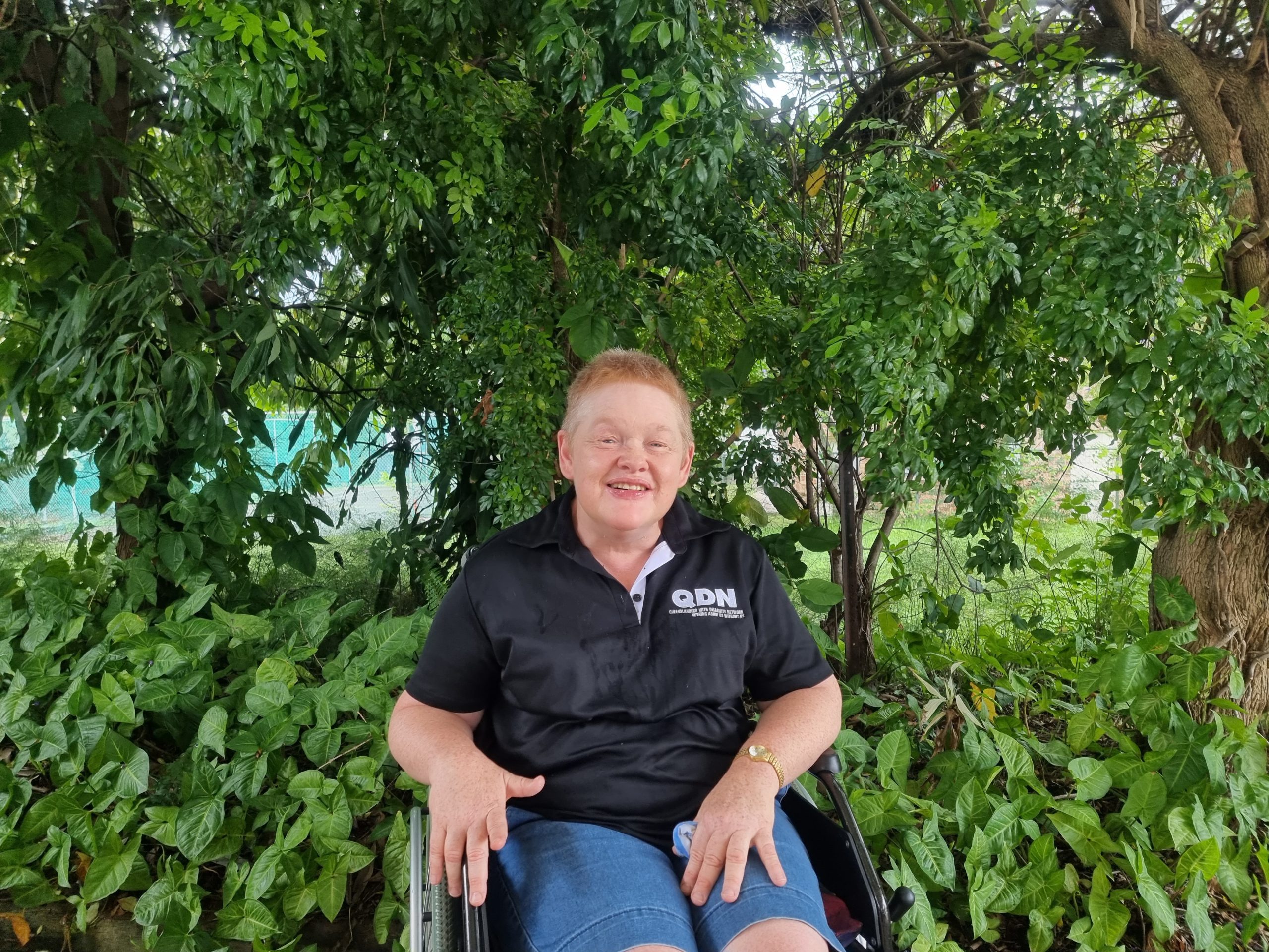 A lady in a wheel chair wearing a QDN polo and sitting in front of a lot of greenery, smiling at the camera.