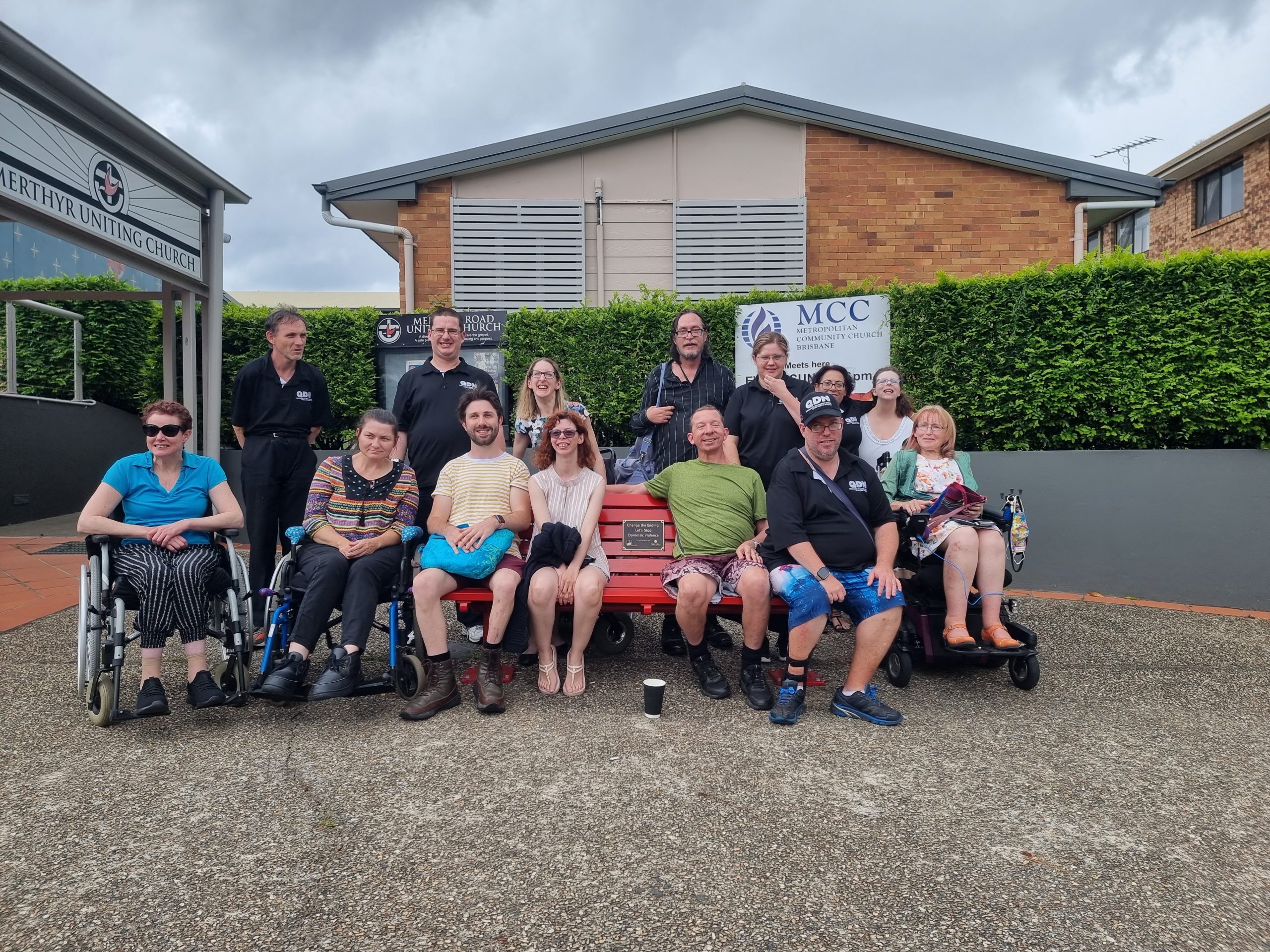 Group of people some in wheel chairs, some sitting on a bench and a row of people standing behind. All smiling and looking at the camera.