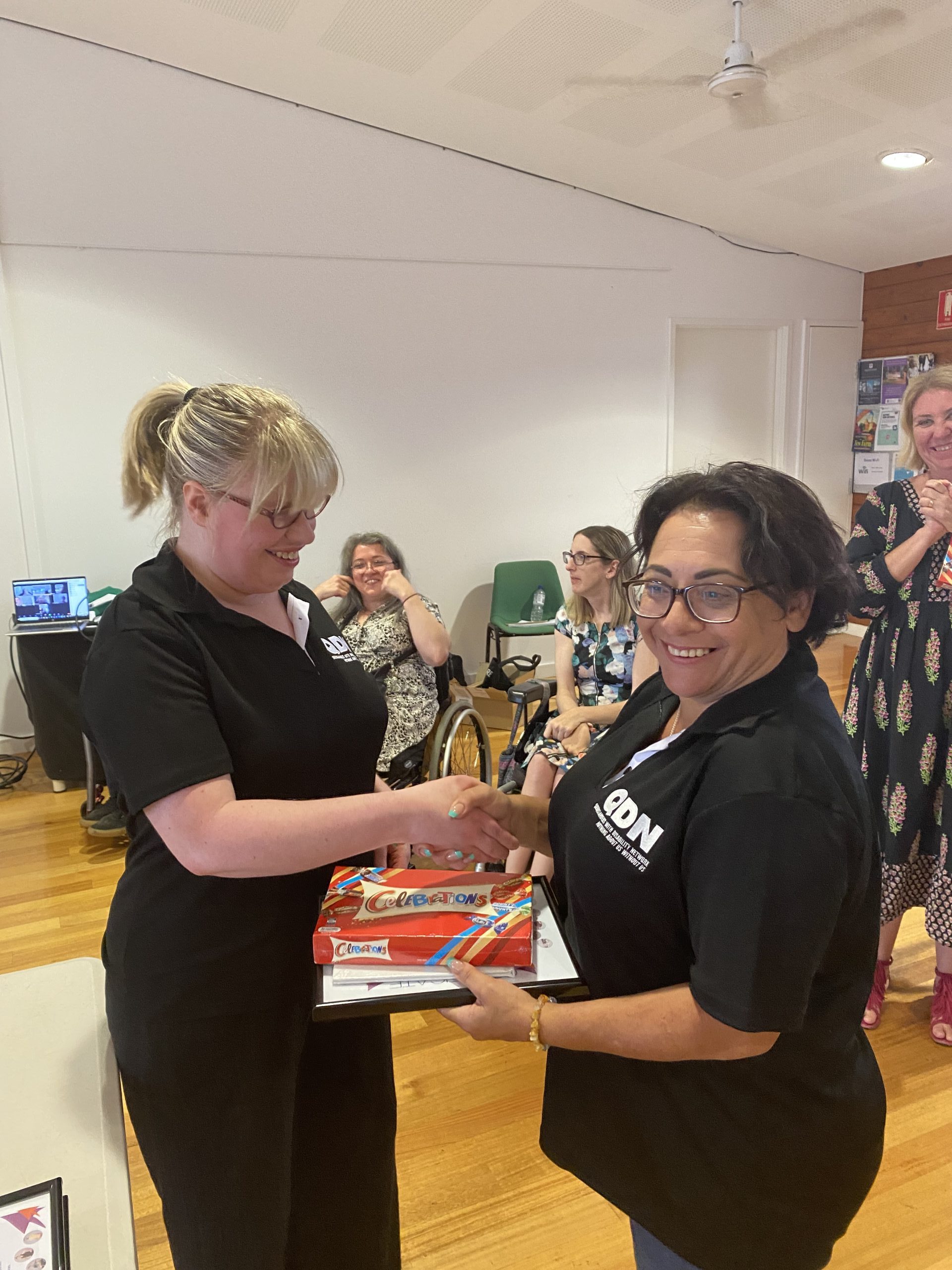 Two woman wearing QDN shirts. They are shaking hands and exchanging a gift and a certificate.