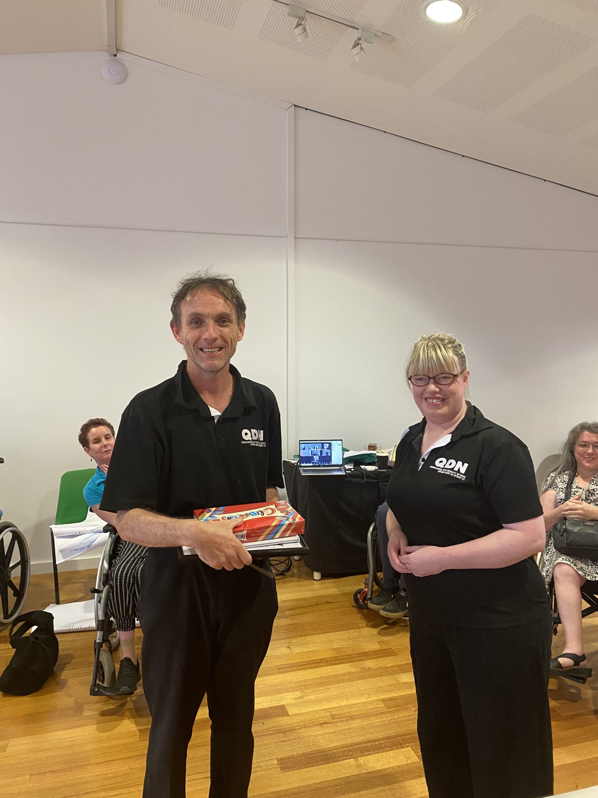 A man and a woman, both in QDN shirts, smiling at the camera. Them an is holding a present and a certificate.
