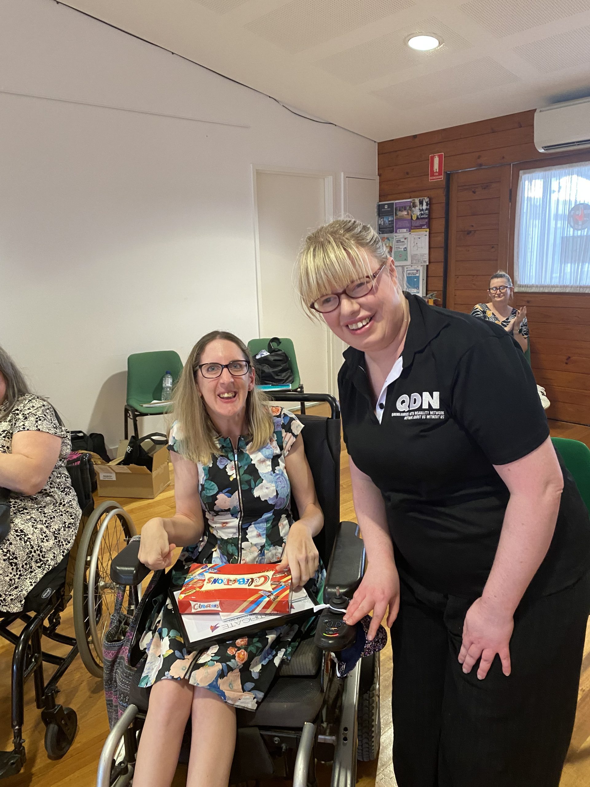 Two women, one sitting in a wheelchair wearing a blue patterned dress and the other lady is standing next to her wearing a QDN shirt.