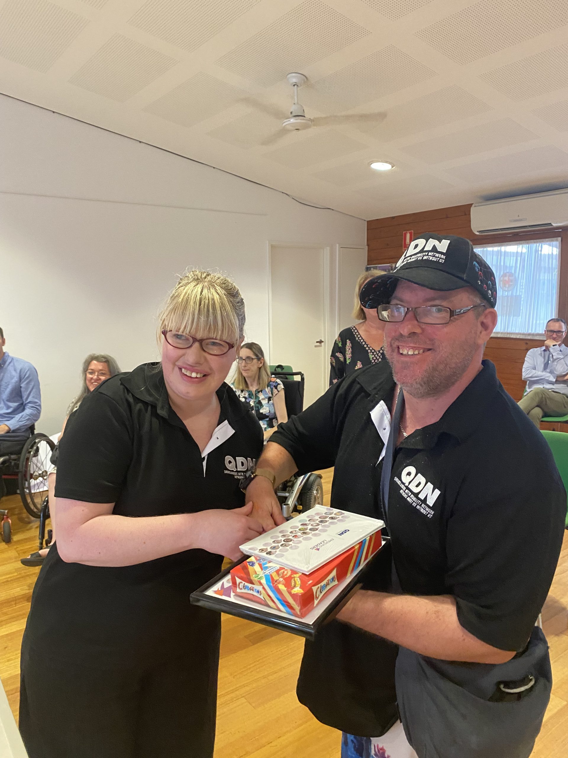 A woman and a man both wearing QDN shirts. They are shaking hands and exchanging a gift and a certificate. They are both looking at the camera smiling.