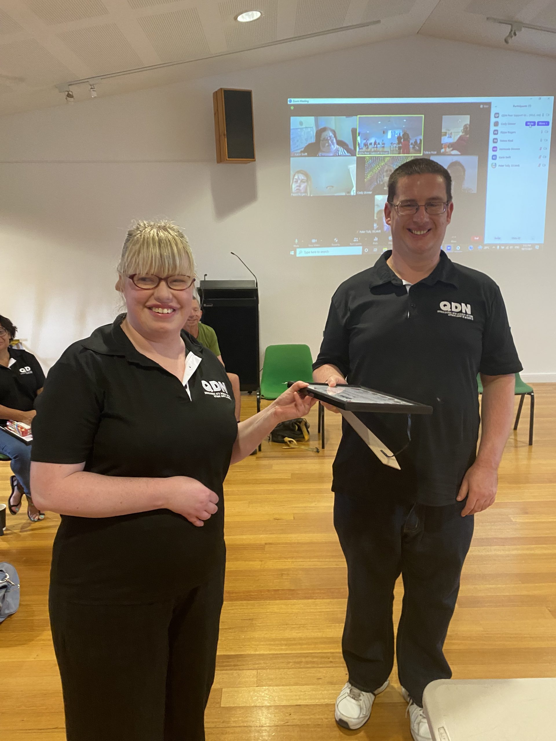 A man and a woman standing next to each and both wearing QDN shirts. The woman is passing the man a certificate and they are both looking at the camera smiling.