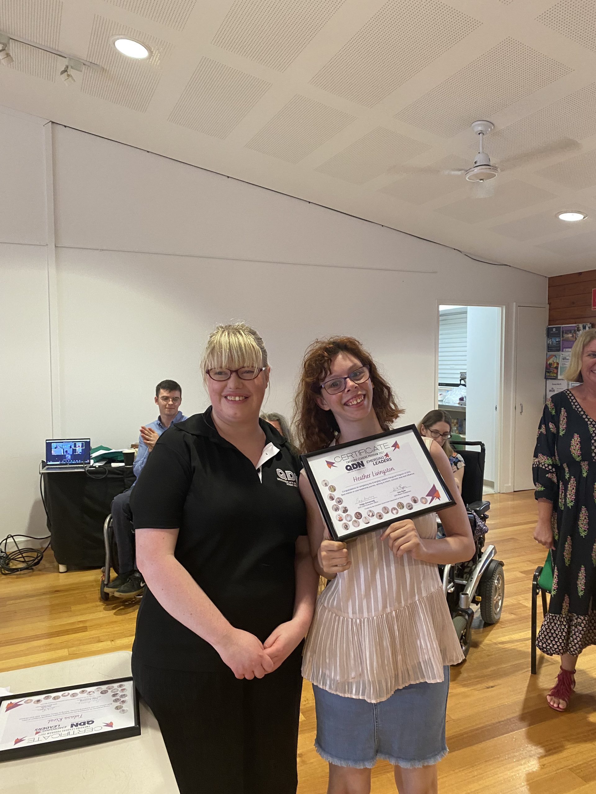 Two ladys standing next to each other smiling at the camera. The lady on the right is holding a framed certificate.