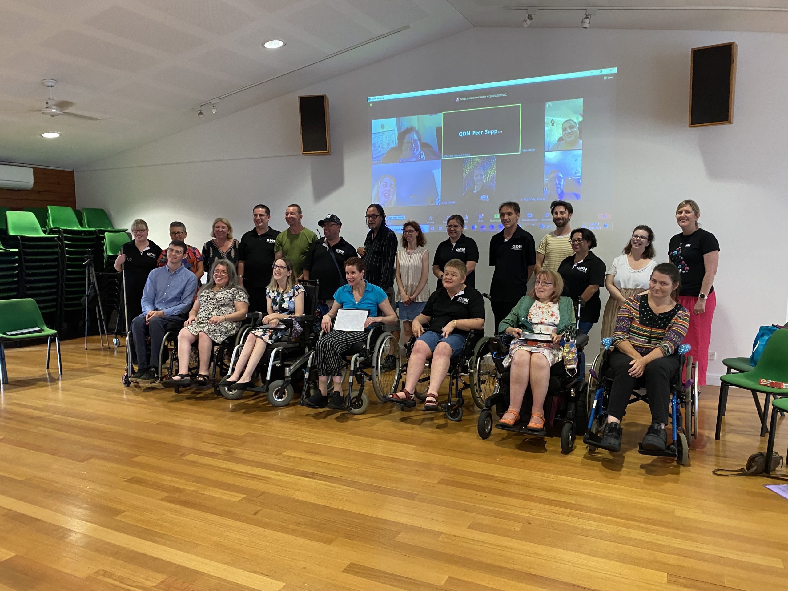 A large group of people in two rows, the front row everyone is sitting in wheelchairs and then a standing row behind them. There is also a projected picture of people on a zoom call on the wall behind them.