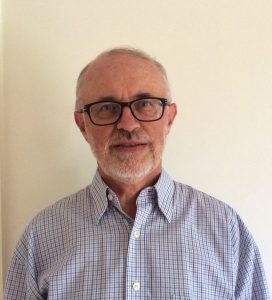 A photo of a man standing in front of a blank wall. He is wearing a business buttoned up shirt and glasses.