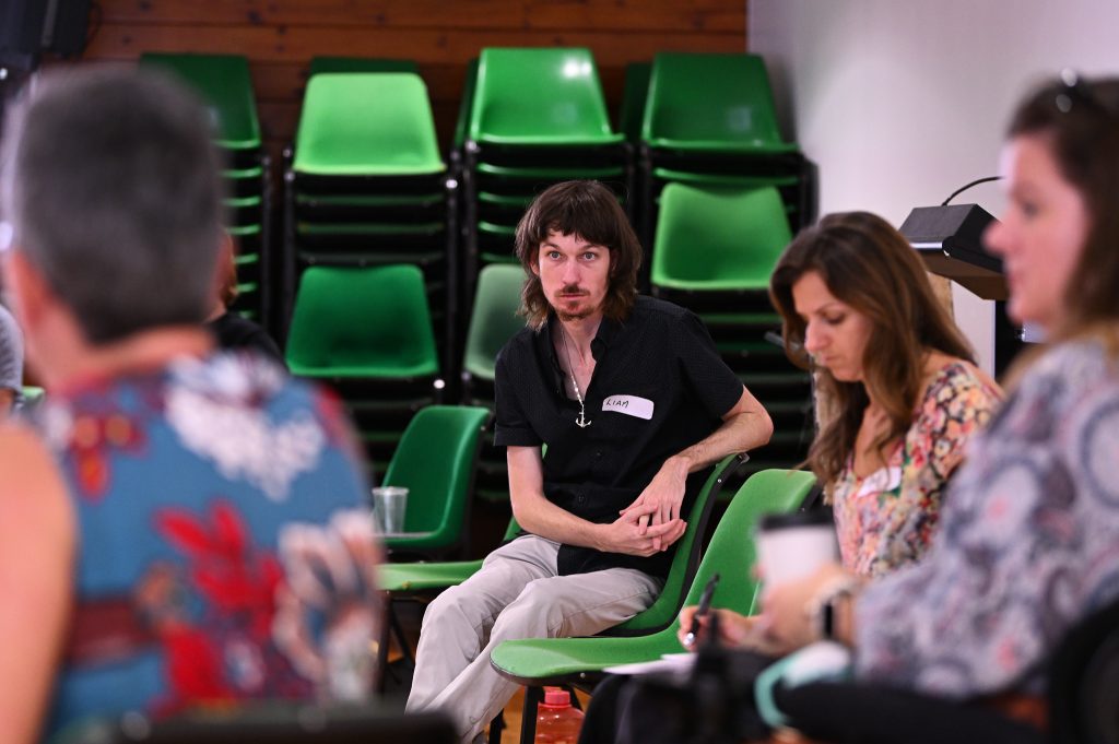 People sitting on chairs in a circle, the photo focuses on one man with brown hair wearing a black polo.