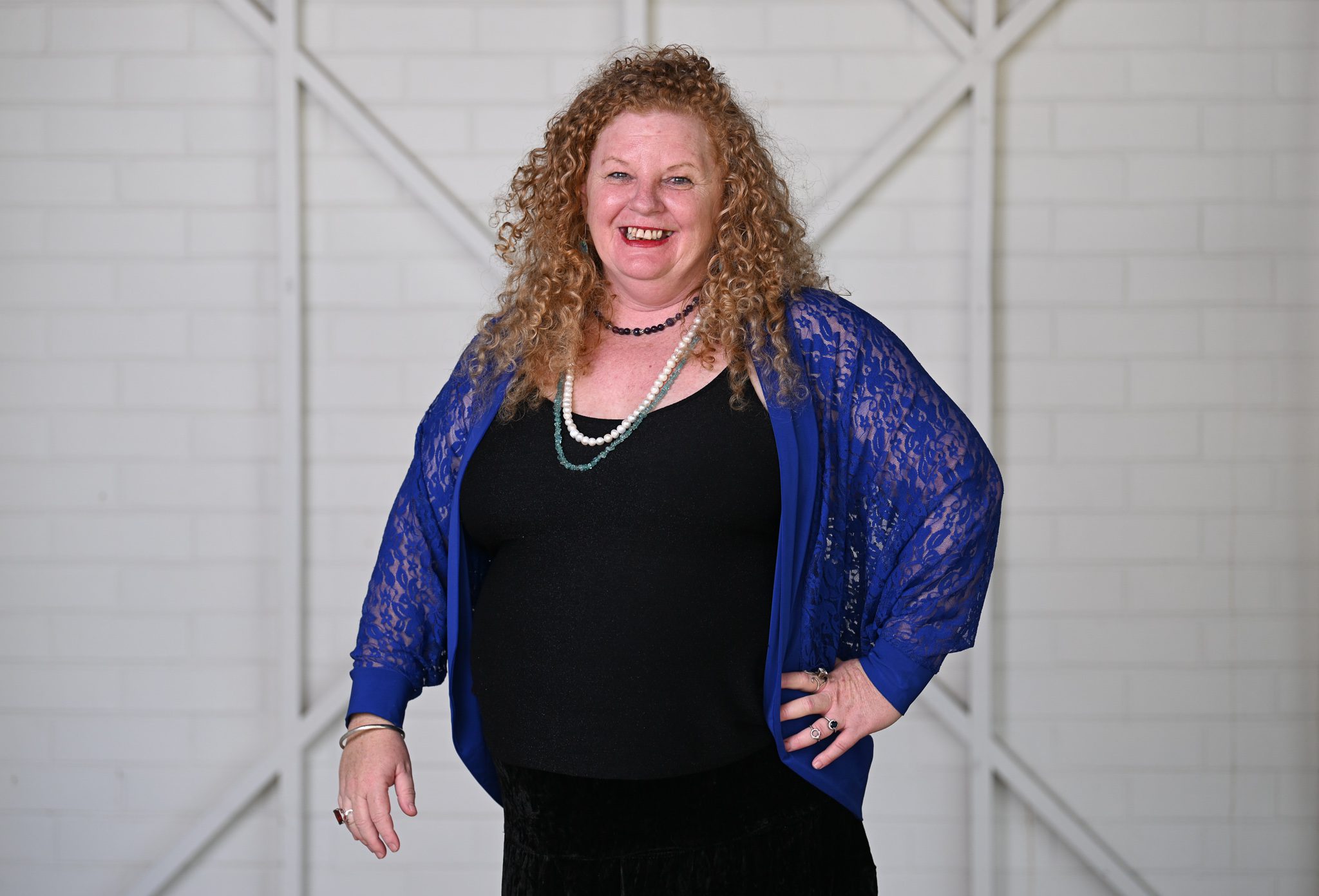 A woman with light curly long hair, wearing a black shirt with a blue cardigan, smiling at the camera.