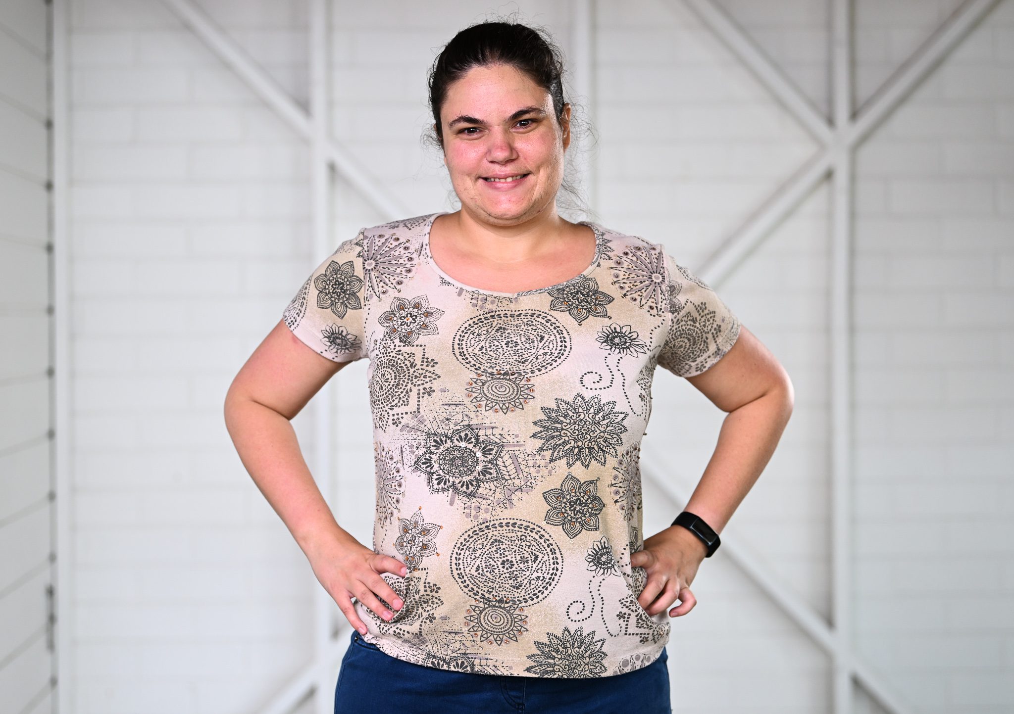 A girl with dark brown hair pulled back, wearing a patterned shirt, with her hands on her hips smiling at the camera.