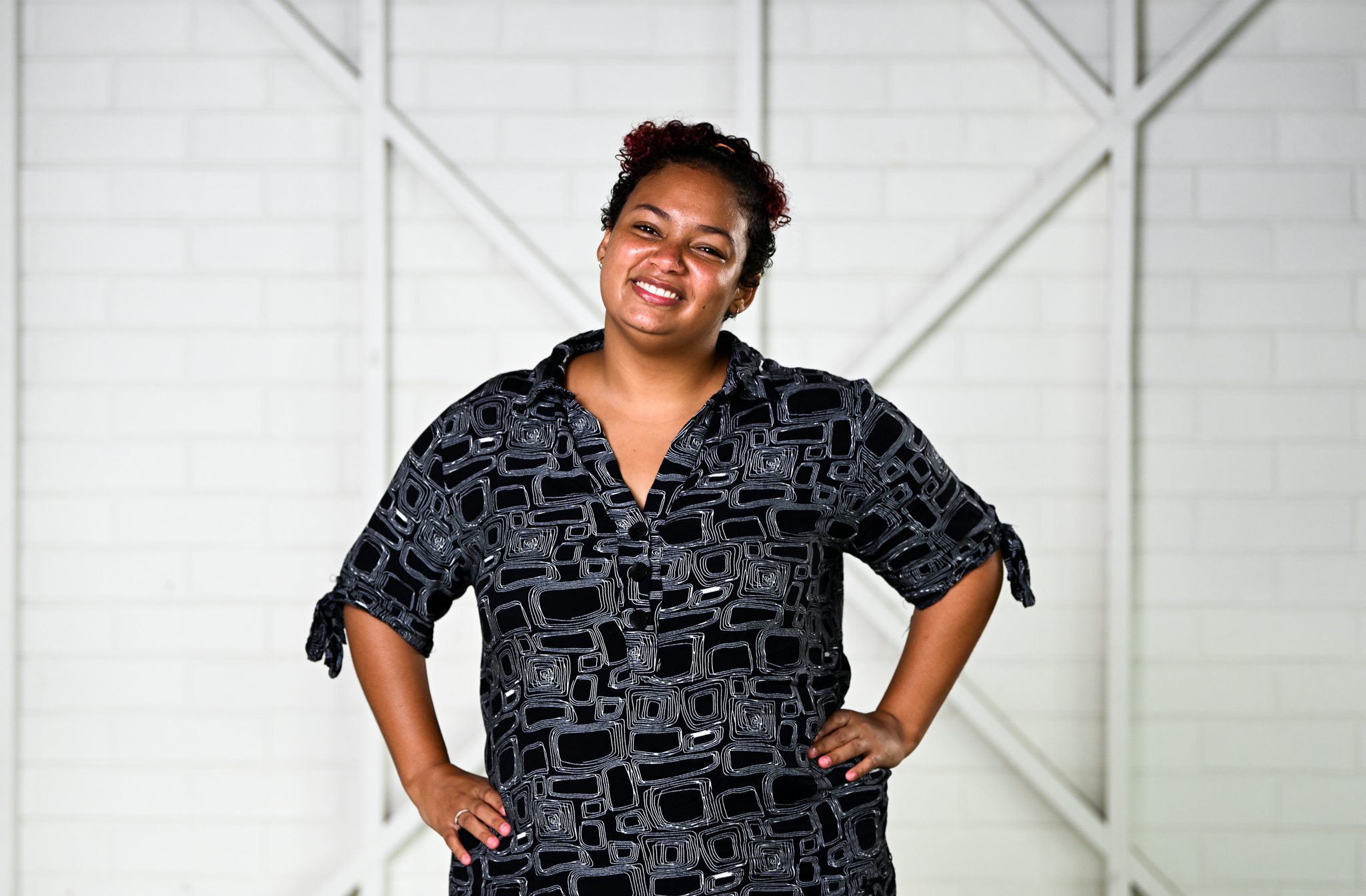 A woman with dark brown short hair, wearing a black and white patterned buttoned up shirt, with her hands on her hips and smiling at the camera.