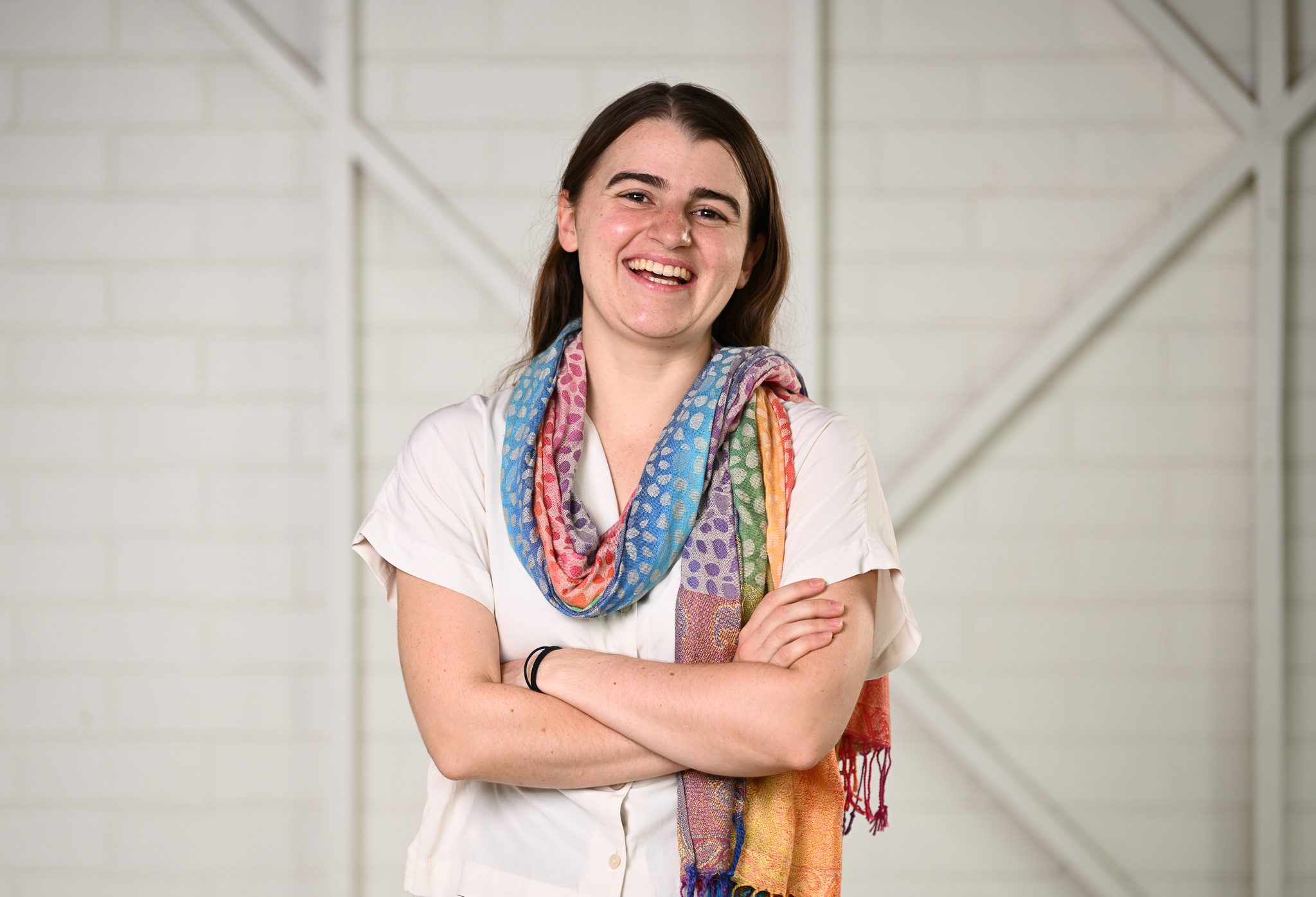A girl with long brown hair, wearing a white shirt and colourful scarf, she has her arms crossed and smiling at the camera.