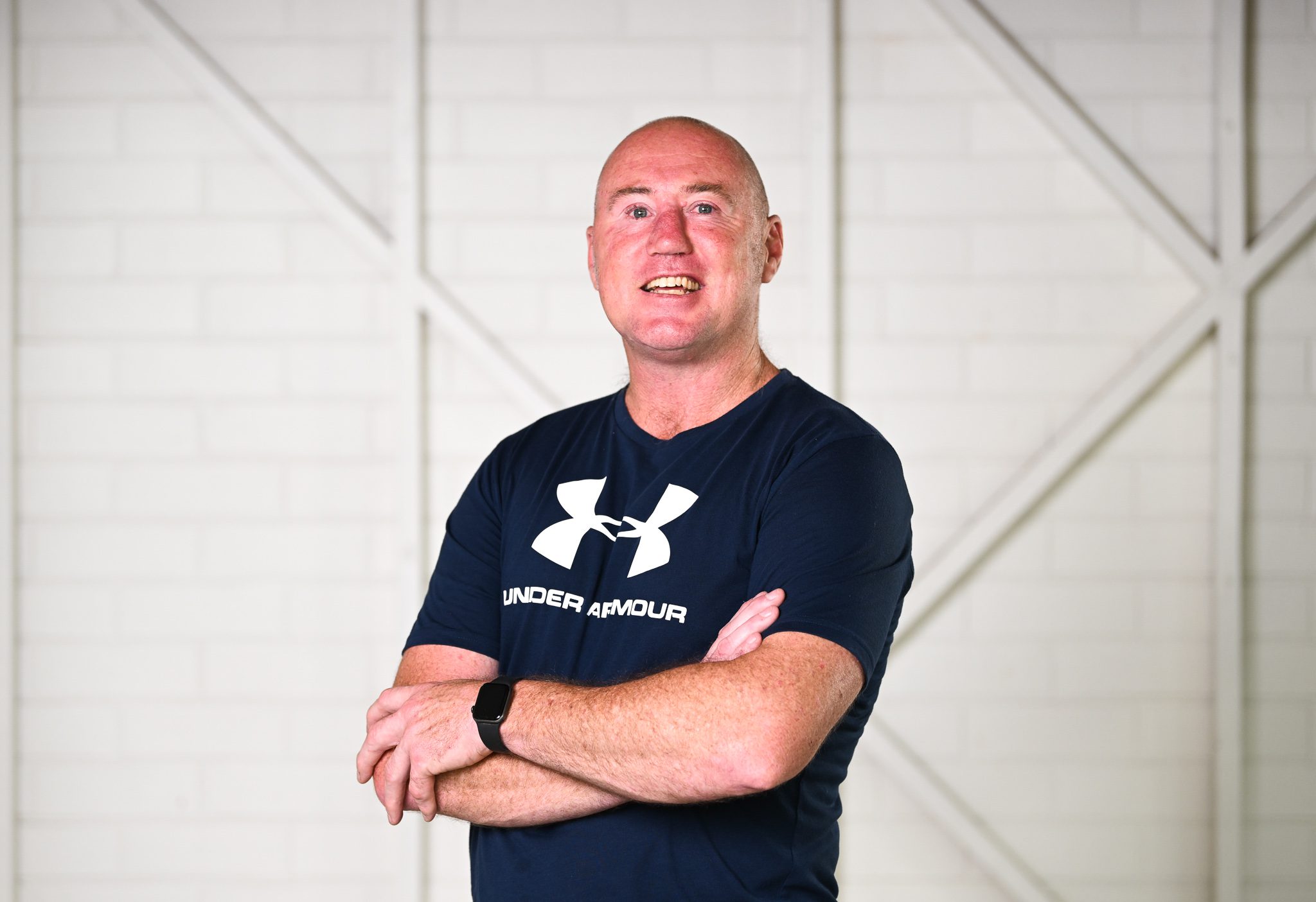 A man with a shaved head, wearing a black tshirt, he has his arms crossed and is smiling at the camera.