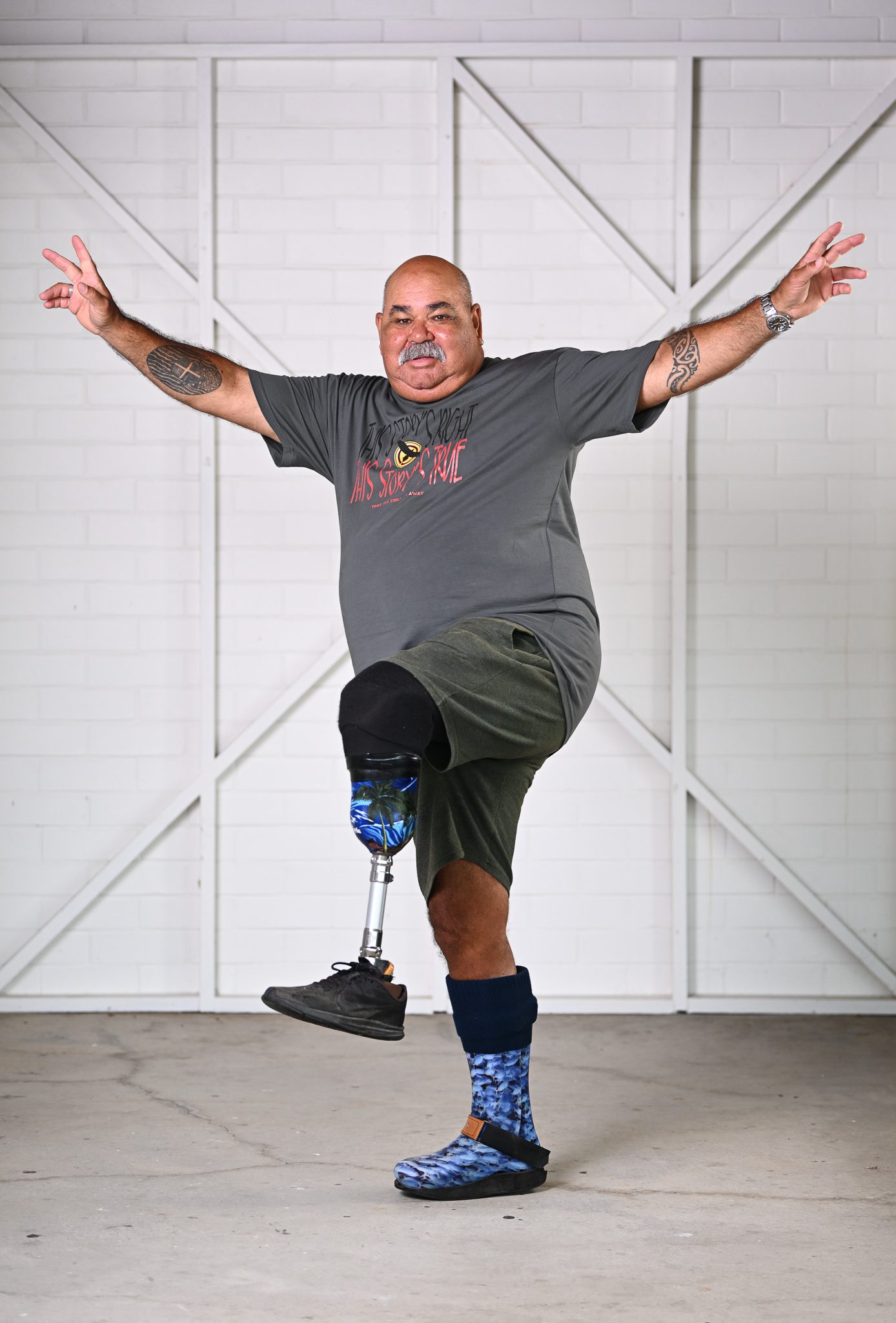 A man with a mustache, wearing a grey tshirt, he has his arms up in the air and is smiling at the camera.