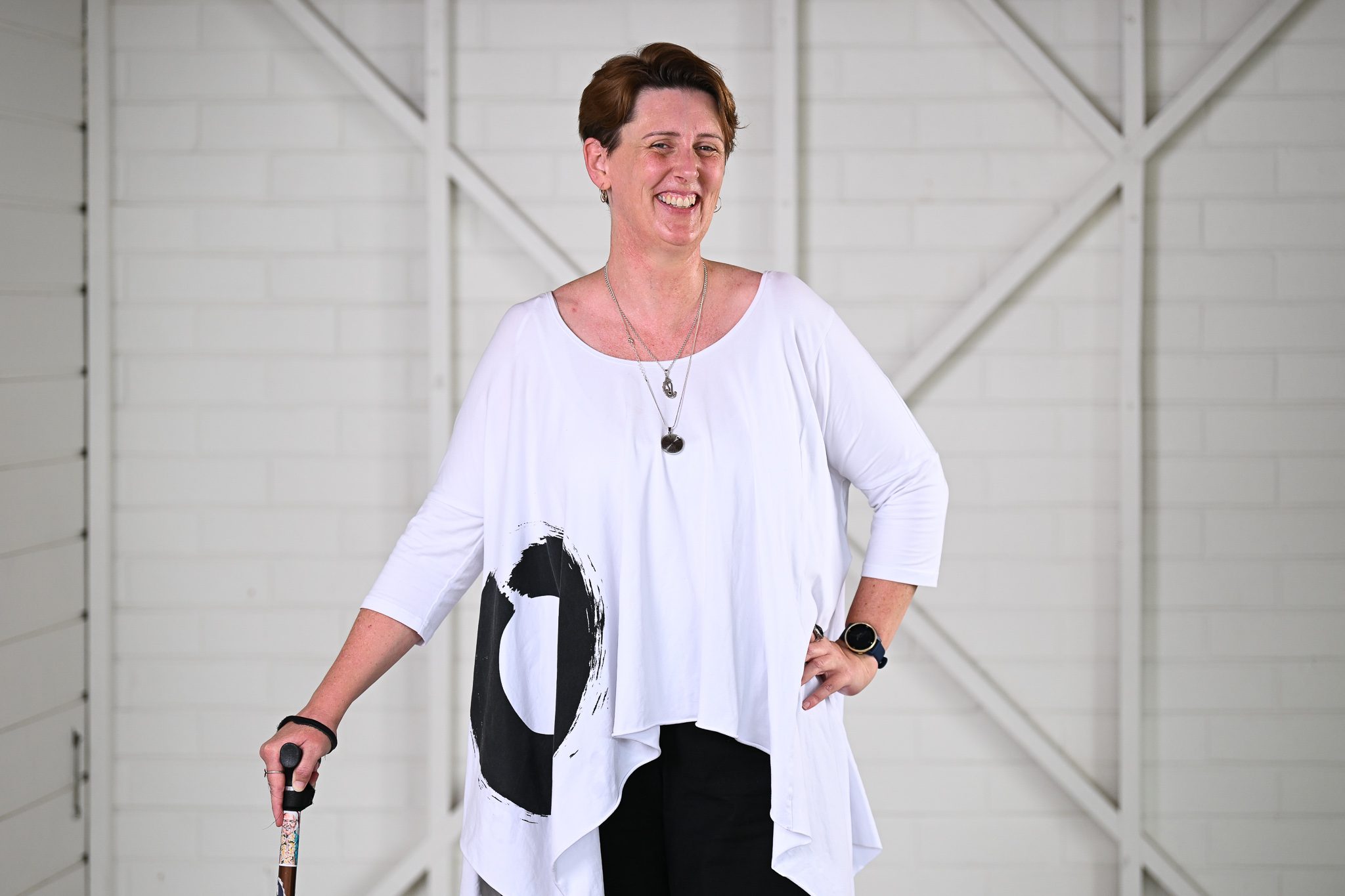 A woman with short brown hair, wearing a white flowing blouse and is holding a walking stick and smiling at the camera.